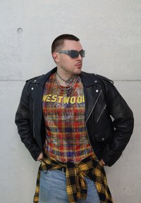 Svart skinn bikerjakke, flerfarget rutete skjorte med "WESTWOOD", gul rutete skjorte knyttet rundt livet, sølv solbriller, lag på lag med kjeder.