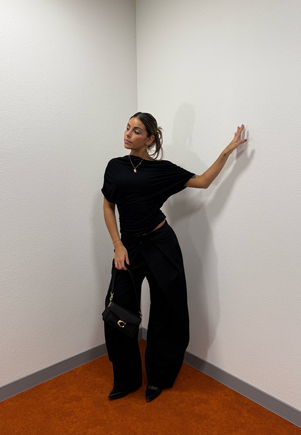 Femme en tenue noire et chaussures pointues se tient dans un coin, tenant une petite pochette noire, une main touchant le mur.