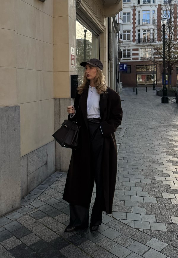 Femme en long manteau sombre et casquette tenant un sac à main, debout sur un trottoir en ville près d'un bâtiment avec des fenêtres en verre.
