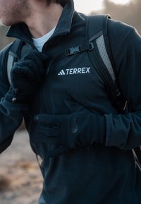 Veste polaire noire avec le logo « TERREX », associée à des gants noirs arborant le logo « The North Face ». Des bretelles de sac à dos visibles sur l'épaule.
