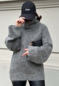 A person dressed in an oversized grey fuzzy sweater and black leather trousers, holding a black clutch and adjusting a black cap featuring the text "MY DOG IS MY CHILD."