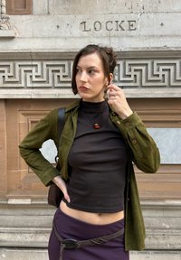 Groene suède knoopjack, zwarte coltrui, paarse rok en een hangerketting. Kenmerkt zich door textuurstof en een casual pasvorm.