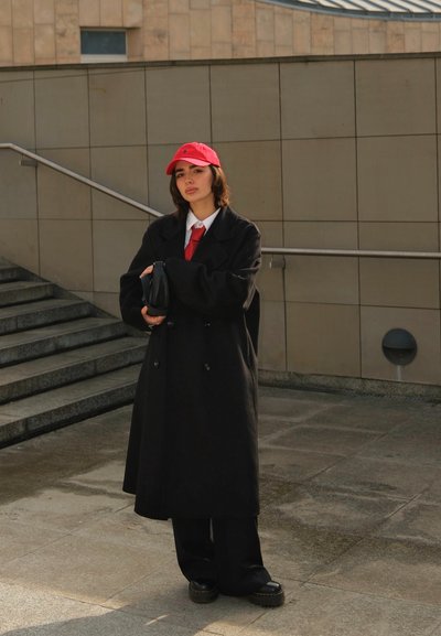 Abrigo negro de doble botonadura, camisa blanca de botones, corbata roja y gorra roja; sosteniendo un pequeño bolso negro; pantalones oscuros de pierna ancha y zapatos de suela gruesa.