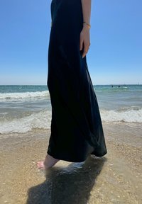 Černé, splývavé maxi šaty ke kotníkům, nošené na písčité pláži. Naboso v mělké vodě, s jasně modrou oblohou a oceánem v pozadí.