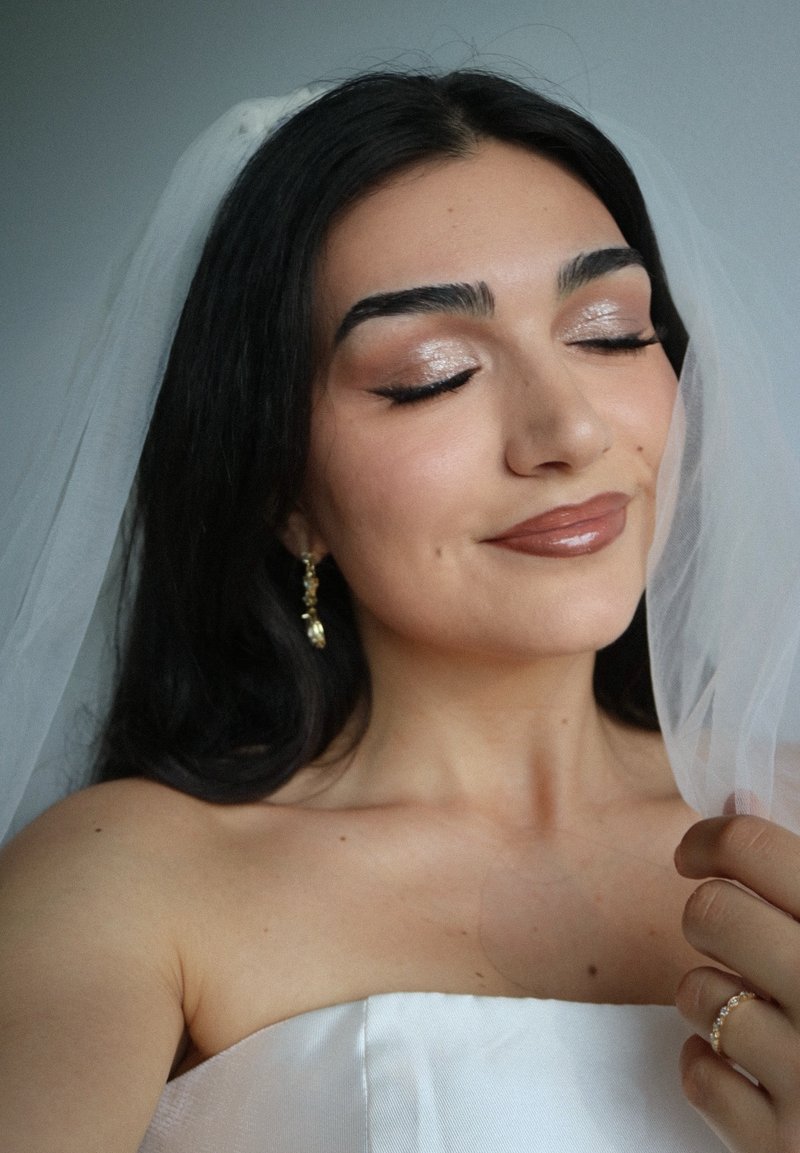 Femme aux cheveux foncés et yeux fermés portant une robe blanche bustier, un voile transparent, une boucle d'oreille en or, tenant le voile de la main et montrant une bague.