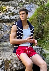 Modelo vestindo uma t-shirt branca, um colete sem mangas listrado em azul marinho e branco, e calções vermelhos com detalhes brancos, sentado em grandes rochas.
