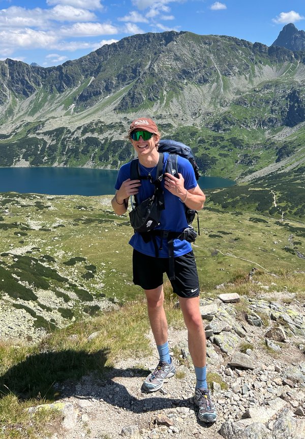 Vandrer i en blå skjorte, sorte shorts og grå vandresko, bærende en blå rygsæk med grønne detaljer mod en bjergagtig baggrund.