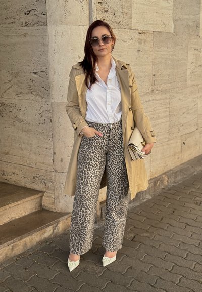 Trench de color beige, camisa blanca con botones, pantalones de leopardo, zapatos blancos de punta, gafas de sol grandes y bolso de mano, de pie sobre una superficie pavimentada.