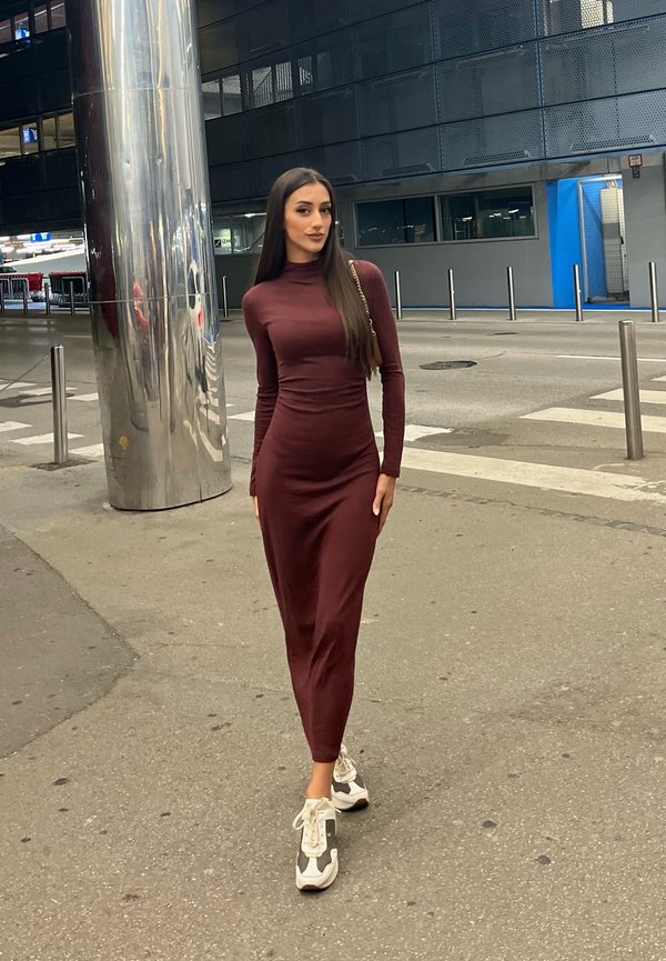 Une femme vêtue d’une robe bordeaux et de baskets blanches pose à l’extérieur d’un bâtiment moderne, à côté d’une colonne métallique réfléchissante.