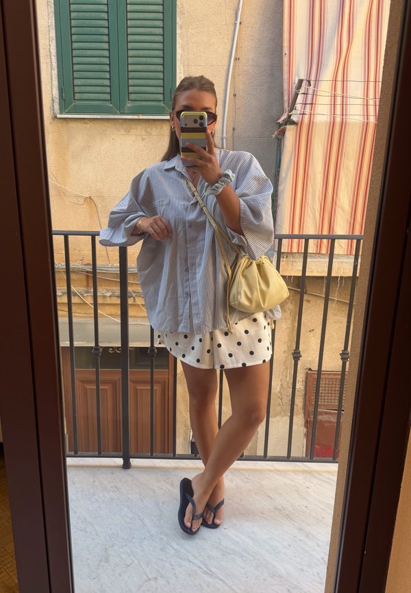 Vrouw met een oversized gestreept overhemd en korte stippenbroek maakt een spiegelselfie op een balkon met een gele tas en slippers.