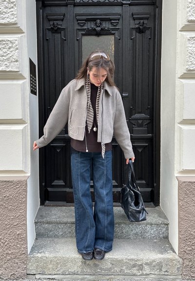 Joven mujer con chaqueta gris, bufanda de rayas y vaqueros anchos, de pie sobre escalones de piedra, sosteniendo un bolso negro, delante de una puerta negra ornamentada.