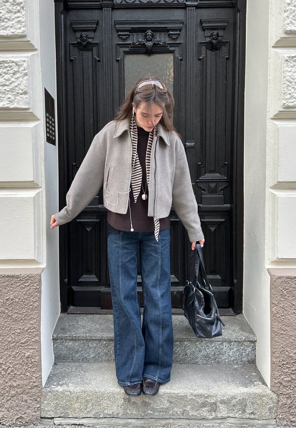 Nuori nainen harmaassa takissa, raidallisessa huivissa ja leveissä farkuissa seisoo kivisillä portailla, pitelee mustaa käsilaukkua mustan koristeellisen oven edessä.