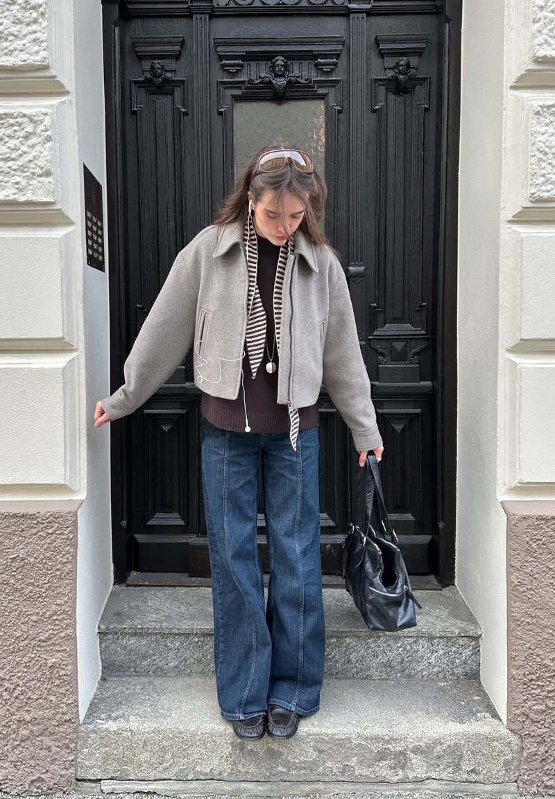 Giovane donna con giacca grigia, sciarpa a righe e jeans larghi, in piedi su gradini di pietra, con una borsa nera davanti a una porta nera ornata.