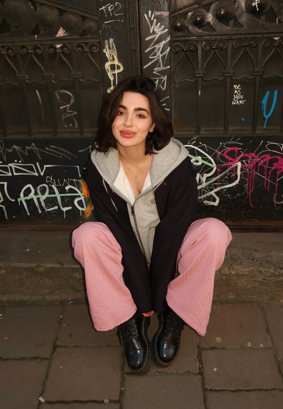 Conjunto casual que presenta una sudadera gris, una chaqueta negra oversize y pantalones de pierna ancha a rayas rojas. Botas negras con cordones. Fondo con graffiti.