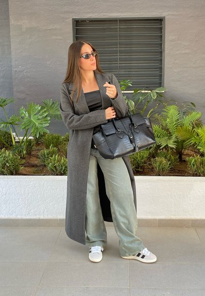 Largo abrigo gris de lana sobre una blusa negra, combinado con jeans de pierna ancha verdes claros y zapatillas blancas. Un bolso cruzado negro de cocodrilo acentúa el conjunto.