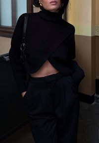 Suéter negro de canalé con un cuello alto, que presenta un corte asimétrico, combinado con pantalones negros de talle alto. Pendientes circulares dorados.