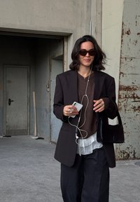 Sieviete, tērpta tumšā žaketē, brūnā džempera, slāņotā baltā blūzē, melnās biksēs, saulesbrillēs, turēdama telefonu ar austiņām un melnu roku somu industriālā vidē.