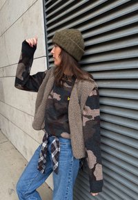 Brun og grå kamuflage-sweater lagdelt med en tan fleecevest, blå jeans og en grøn strikket hue. Sort og hvid ternet skjorte bundet om livet.