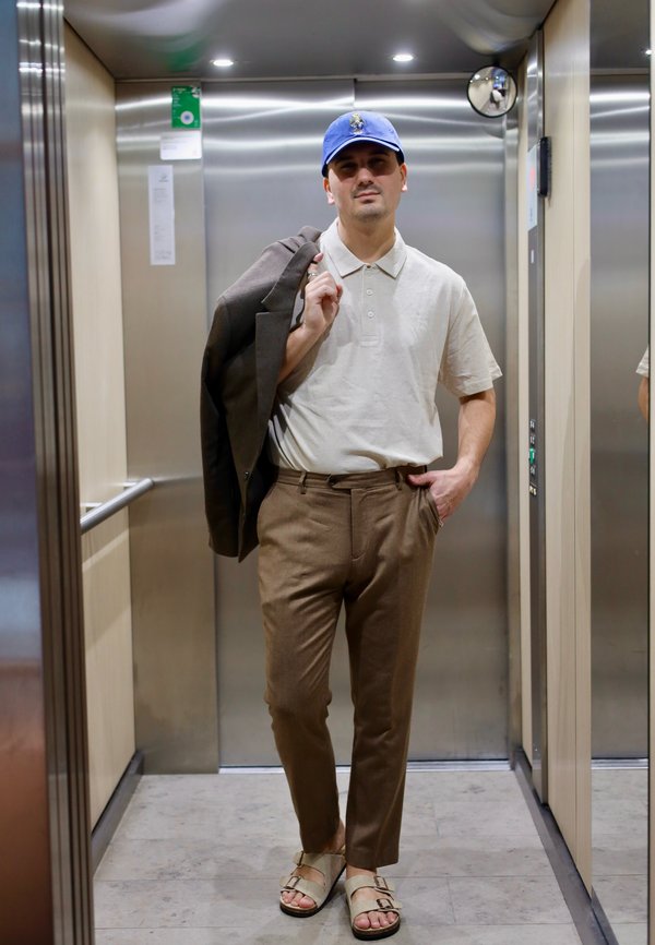 Mand iført blå kasket, hvid polo, brune bukser og sandaler, bærende en jakke over skulderen inde i en metal elevator.