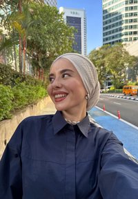Lachende vrouw met een beige hoofddoek en een marineblauwe shirt, staande op een stadsstoep met groene planten en hoge gebouwen op de achtergrond.