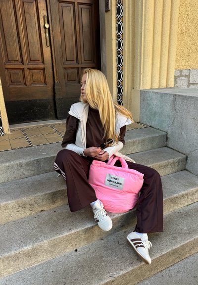 Bolsa aislante rosa con asas superiores, mostrando una etiqueta. Conjunto informal marrón con zapatillas blancas y marrones. Sentado en escalones de piedra.