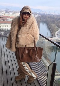 Personne portant un manteau en fourrure beige, des lunettes de soleil et une casquette beige, tenant un sac à main marron, debout sur un balcon avec vue sur la rivière et le paysage urbain en hiver.