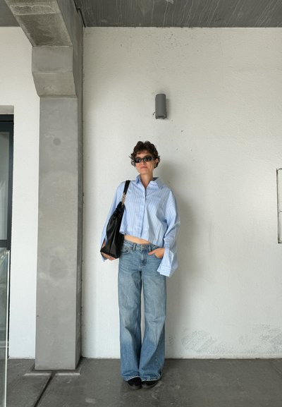 Camisa corta a rayas azul y blanca, pantalones vaqueros de denim claro y holgados, bolso negro de hombro y zapatos oscuros, frente a una pared blanca lisa.