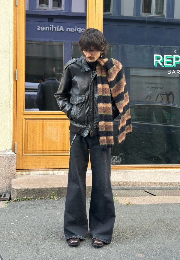 Black leather jacket with front pockets, wide black pants, and a striped brown and black scarf. Circular earrings and brown shoes.