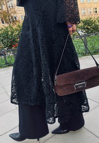Czarna koronkowa sukienka maxi z długimi rękawami, nałożona na czarne spodnie, w zestawie z ciemnobrązową zamszową torebką z srebrnymi akcentami.