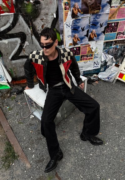Persona con gafas de sol y una chaqueta a cuadros negra, roja y blanca sentada en una caja blanca junto a una pared cubierta de grafitis y carteles.