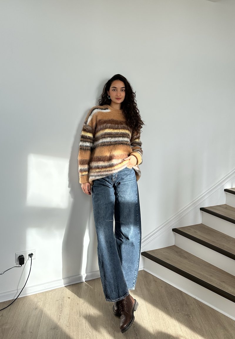 Woman with long curly hair wears a striped fluffy jumper, wide-leg jeans, and brown boots, standing near stairs in a sunlit room.