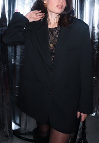 Μαύρο oversize σακάκι με πέτο και δύο μπροστινές τσέπες, τοποθετημένο πάνω από μια διαφανή μαύρη δαντελένια μπλούζα, με γυαλιστερά κουμπιά και υφή. 