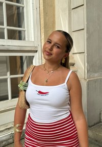 Top a righe bianche con testo "HOT AND SPICY" e grafica di un peperoncino rosso, abbinato a una gonna a strisce rosse e bianche. Gli accessori includono collane sovrapposte e una borsa di paglia.