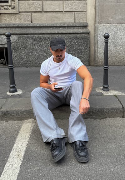 Hombre sentado en un bordillo de piedra, vistiendo una camiseta blanca, pantalones anchos grises y zapatos negros con borlas, utilizando un teléfono móvil. Gorro negro y joyas visibles.