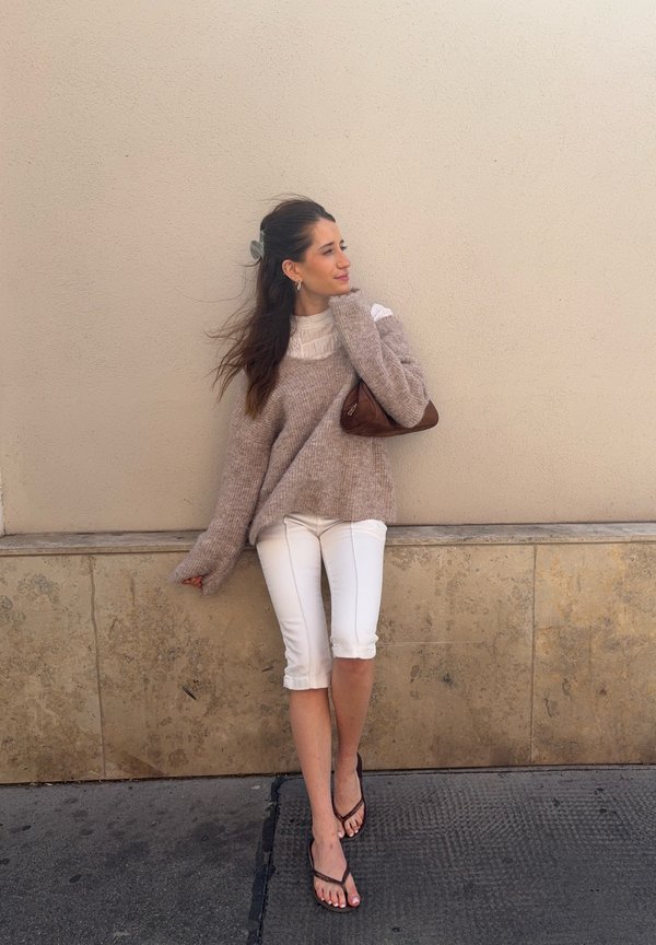 Mujer con un suéter beige, pantalones cortos blancos y sandalias, de pie contra una pared beige sosteniendo un pequeño bolso marrón.