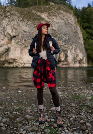 Chaqueta de plumas negra, suéter beige, camisa de franela a cuadros rojos atada a la cintura, leggings marrones y botas de senderismo con una gorra roja. Fondo de río.