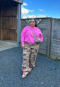 Felpa rosa con logo Nike, pantaloni cargo mimetici, borsa a tracolla beige e sneakers con rifiniture oro e lacci rosa, posizionati su uno sfondo di legno.