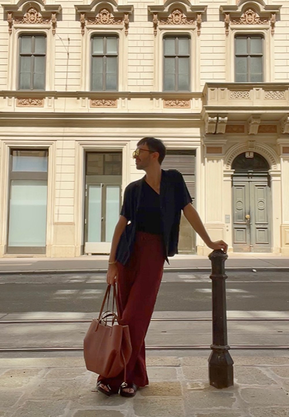 Camisetta nera a maniche corte, pantaloni larghi bordeaux, borsa a mano in pelle marrone; in piedi accanto a un palo decorativo di fronte a un edificio classico.