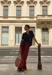 Camisetta nera a maniche corte, pantaloni larghi bordeaux, borsa a mano in pelle marrone; in piedi accanto a un palo decorativo di fronte a un edificio classico.
