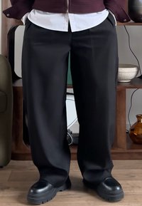 Pantalon noir ample en tissu lisse, taille haute et jambes droites. Associé à des chaussures noires épaisses.