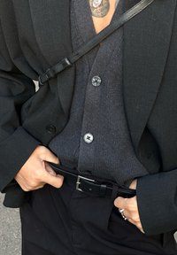 Μαύρο σακάκι πάνω από σκούρο γκρι ριχτό πουλόβερ με κουμπιά. Φοράει μια μαύρη ζώνη στη μέση, με τα χέρια τοποθετημένα στη ζώνη.