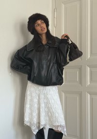 Casaco oversized de couro preto com gola alta, usado sobre um vestido branco de renda. Modelo segura uma pequena mala castanha. Visual texturizado e em camadas.
