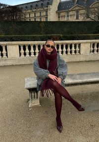 Szary, oversize'owy płaszcz, burgundowy, oversize'owy szalik, burgundowe rajstopy i czółenka z wysmuklonym noskiem. Stoi na kamiennej ławce w ogrodzie.
