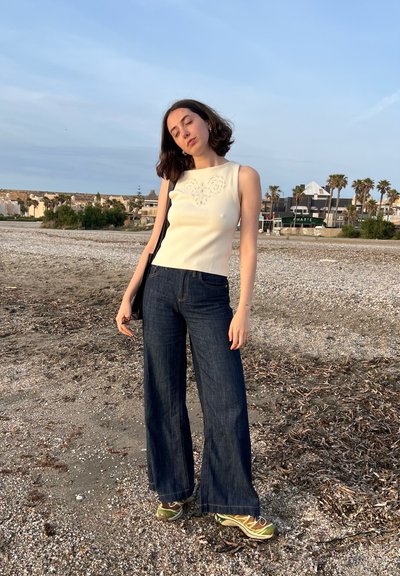 Top sin mangas de color beige claro con detalles bordados, combinado con jeans de mezclilla oscuros de pierna ancha y zapatillas verdosas, situado en una playa de arena.