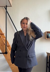 Sacou gri închis, cu două rânduri de nasturi, cu umeri structurați, două buzunare frontale și o textură netedă. Asociat cu o geantă mică neagră.