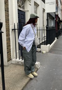Persona che indossa una camicia oversize a righe bianche e blu con pantaloni cargo verdi e scarpe slip-on di colore chiaro, in piedi accanto a un muro di mattoni.