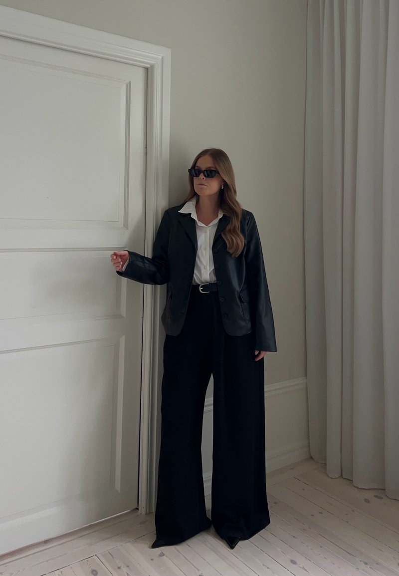 Femme portant des lunettes de soleil, un blazer noir, une chemise blanche et un pantalon large noir debout près d'une porte blanche fermée dans une pièce aux tons neutres.