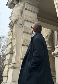 Une personne en long manteau noir se tient à côté d'un pilier en pierre décoratif avec une figure sculptée présentant des textures et motifs détaillés.