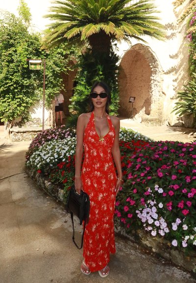 Vestido maxi rojo con estampado floral y diseño de cuello halter, combinado con un bolso negro y sandalias blancas. Rodeada de vibrantes parterres de flores y vegetación.