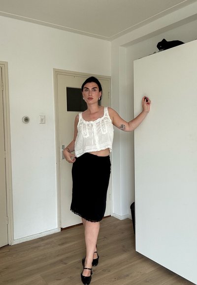 Mujer de cabello oscuro con un top blanco sin mangas y falda negra está descalza sobre suelo de madera, apoyada en un mueble blanco en una habitación blanca.